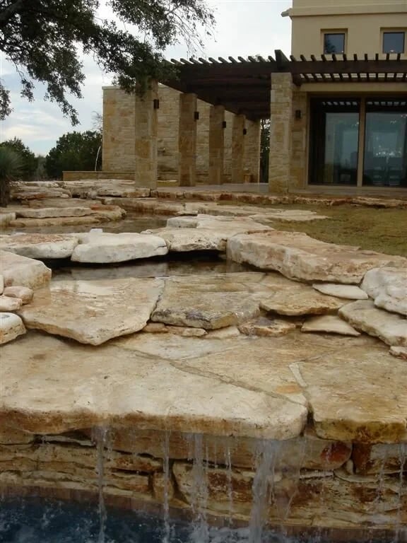 Limestone-slab waterfall spilling into a pool below a stone home with a pergola and glass doors
