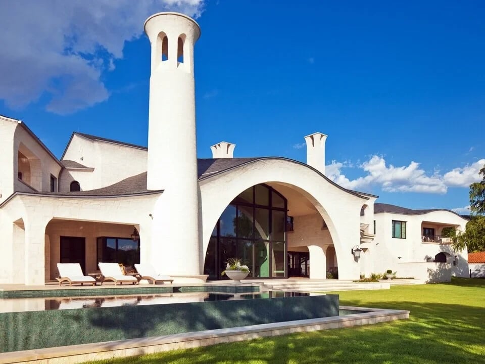 White-stone home with a tall round tower and arched loggia beside a dark geometric pool and lawn