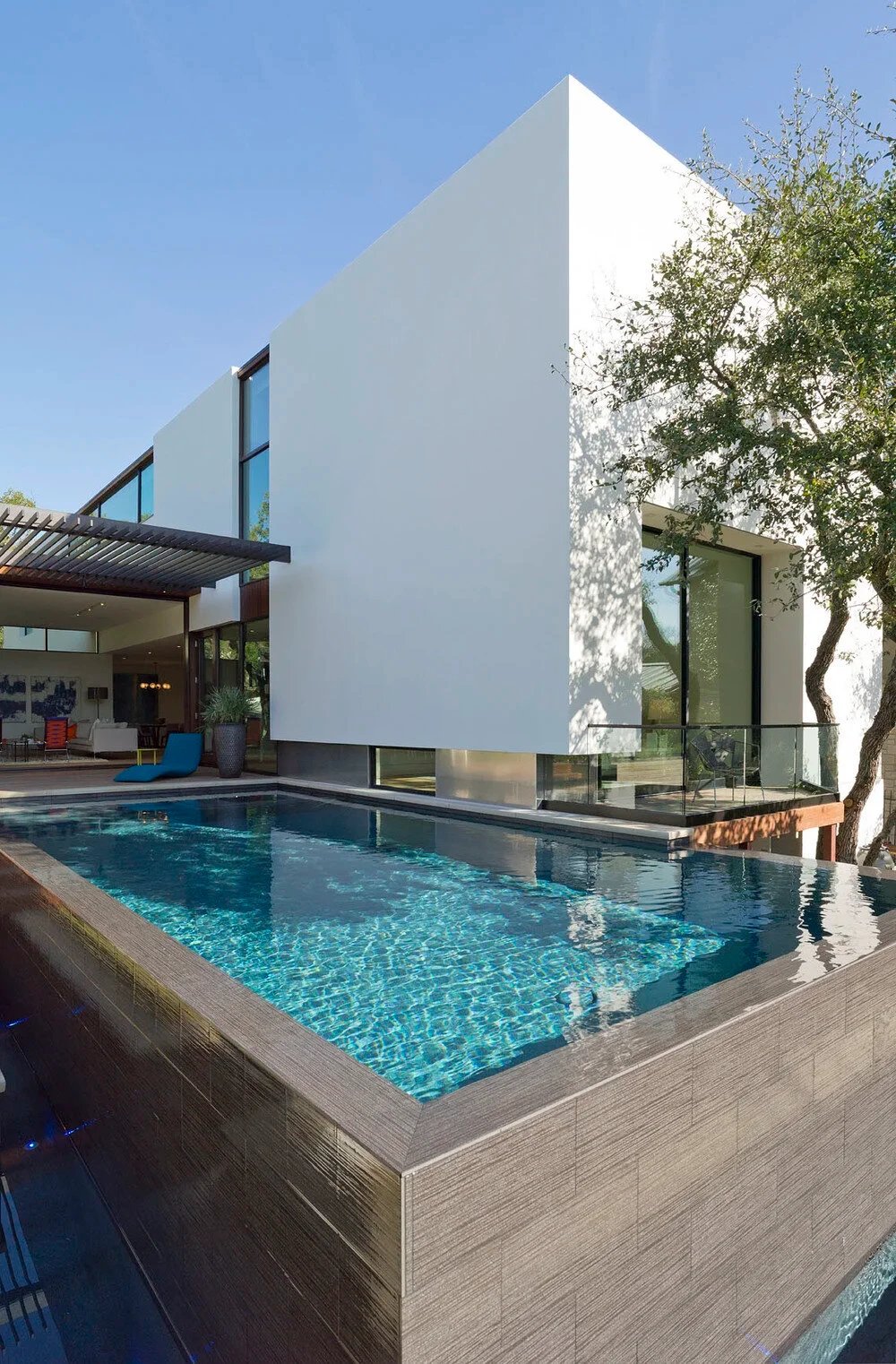 Turquoise infinity pool with wood-clad coping beside a tall white modern home and live oak