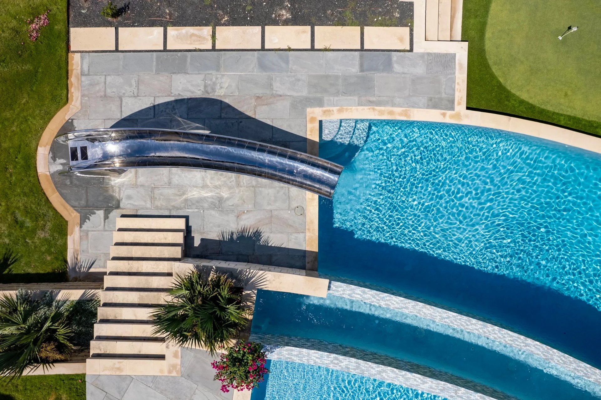 Aerial of a curved stainless waterslide and tiled pool steps, stone deck and putting green below