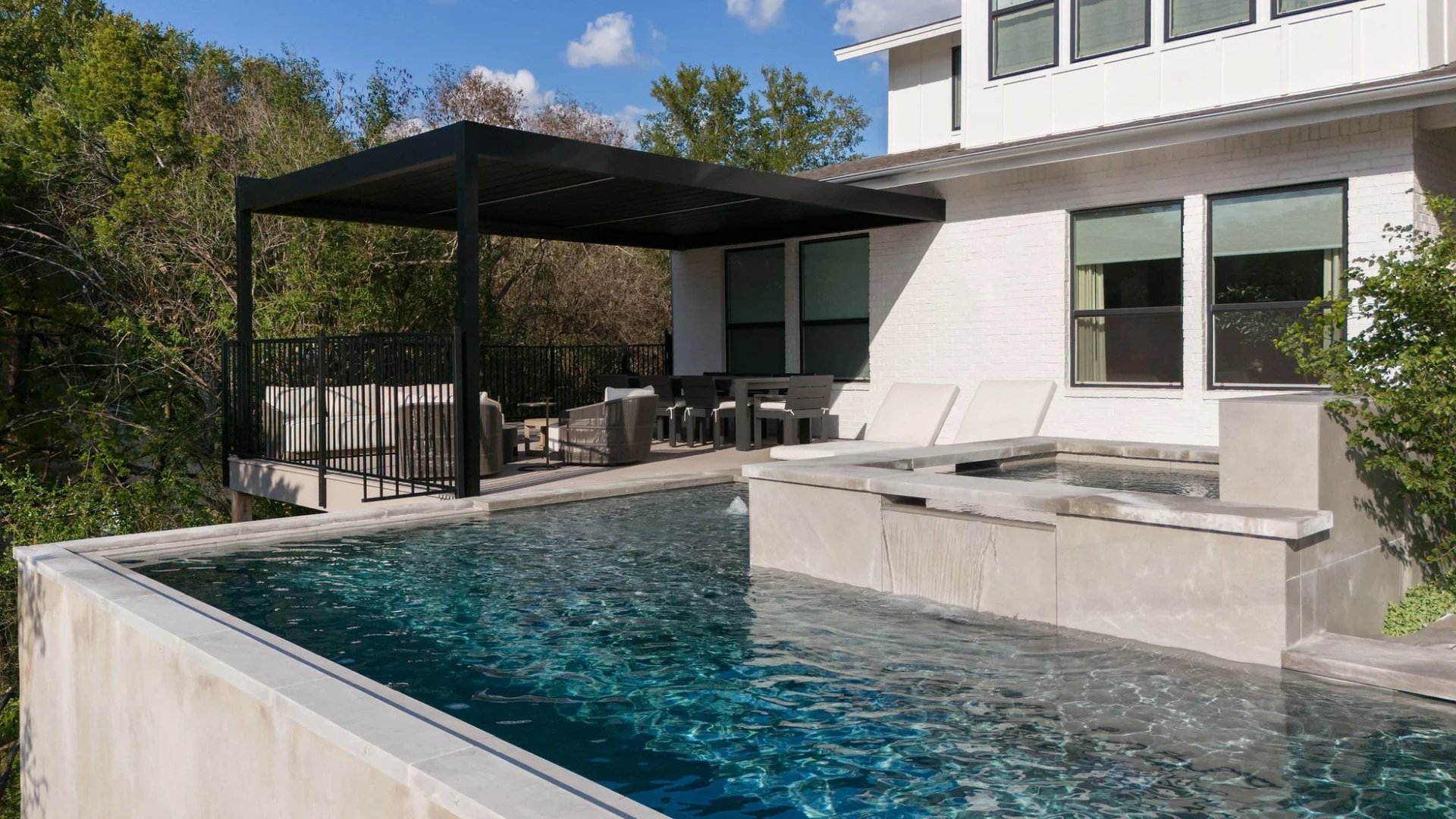 Infinity pool and raised spa beside a white modern home with a black pergola and patio