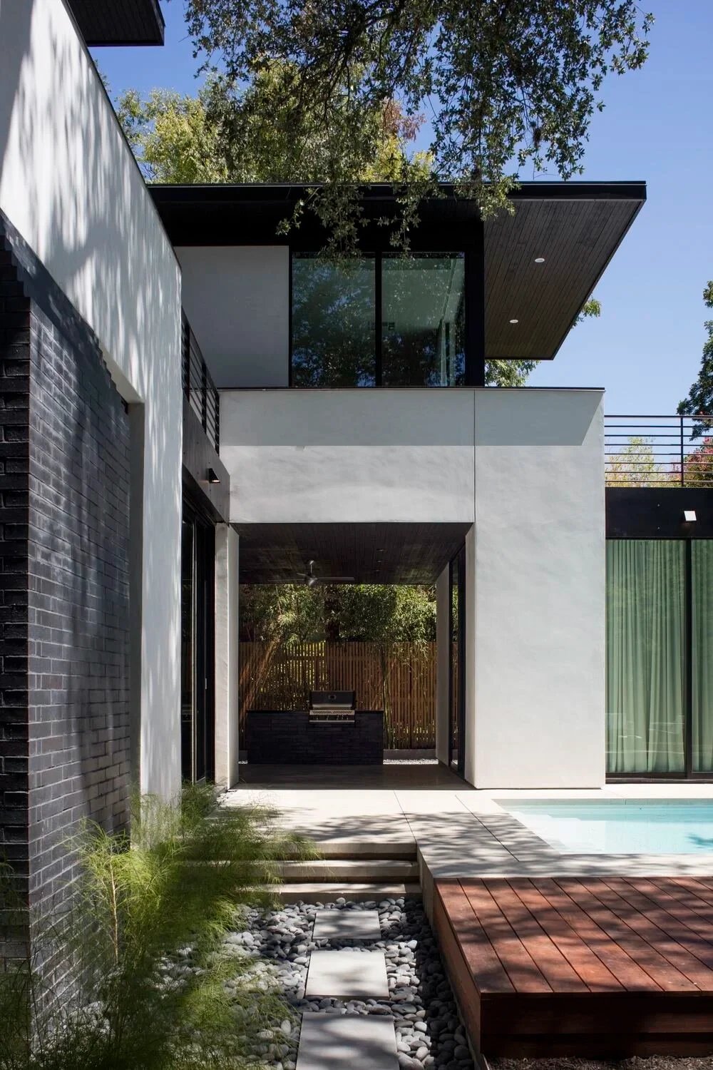 White stucco and dark brick modern home with a stone path and wood deck leading to the pool
