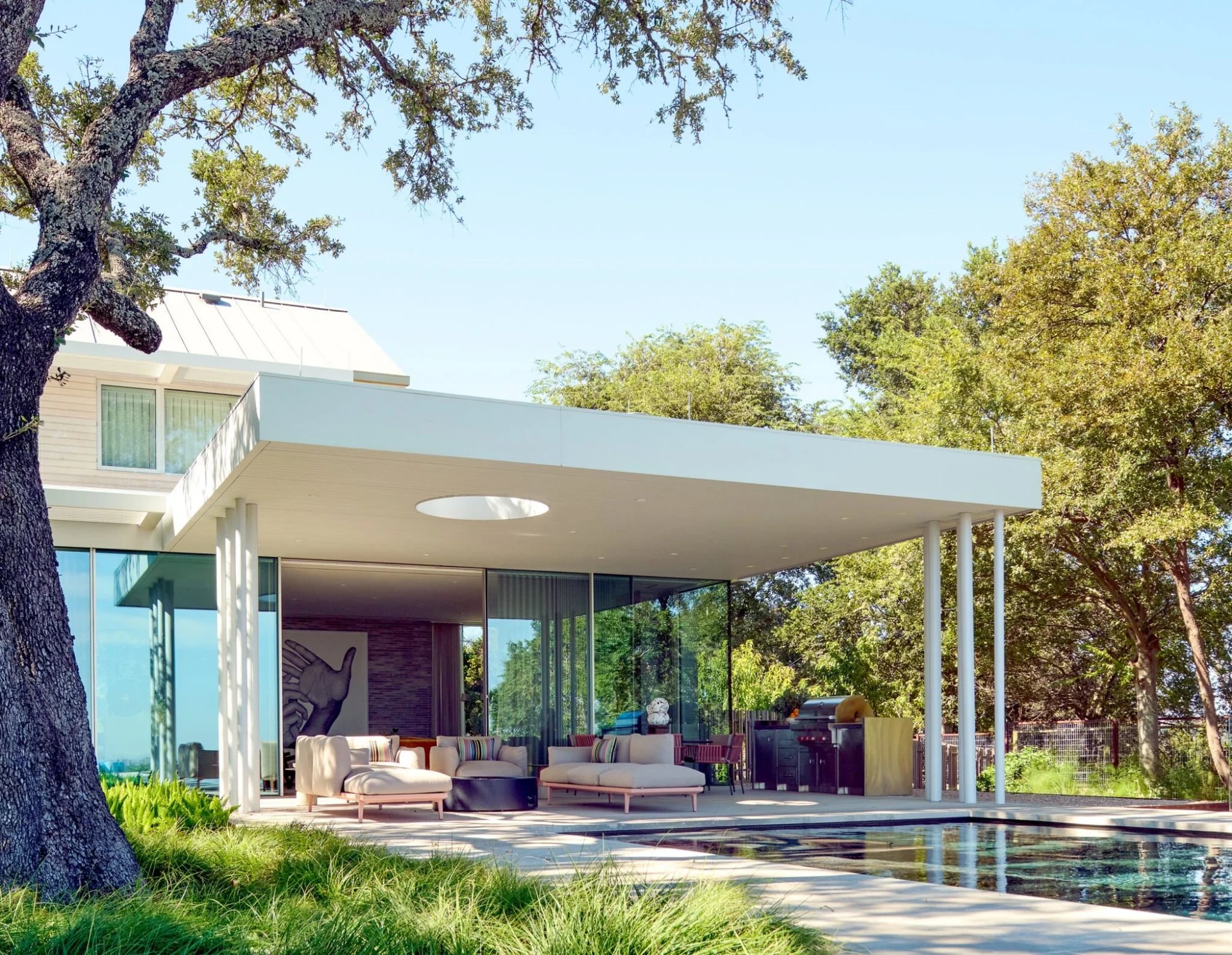 Modern home's flat-roofed glass living pavilion beside a pool, shaded by live oaks over a grassy yard