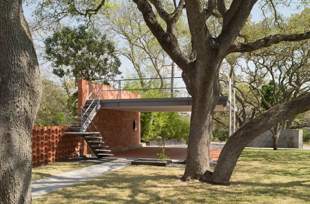 Modern home with a cantilevered upper deck and steel stair, set among live oaks on a green lawn