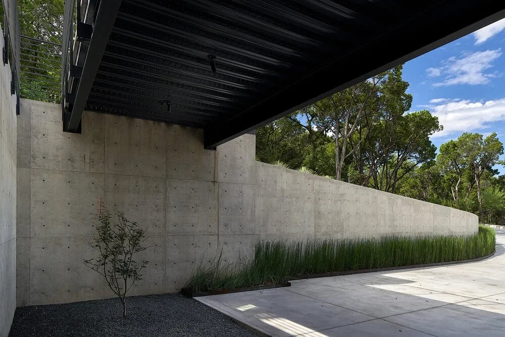Covered concrete walkway along a tall board-formed wall with grasses, opening to woods