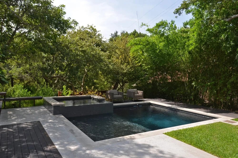 Dark geometric pool and raised spa with stone coping set in a lush, tree-lined backyard