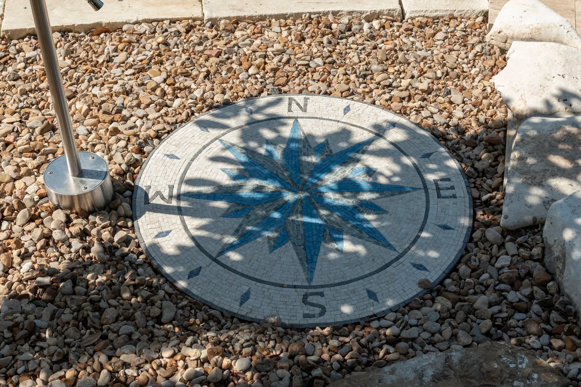 Mosaic-tile compass rose set into a pebble bed beside a pool umbrella pole and limestone