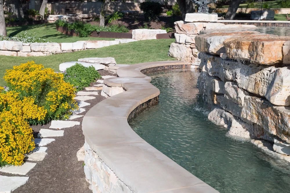 Curving stone deck and waterfall feature beside a spa, with yellow blooms and a manicured lawn