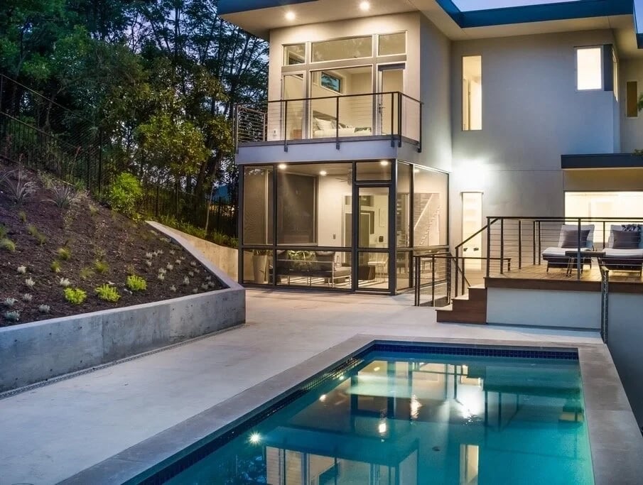 Gunite pool with dark tile reflecting a lit modern home and balcony in Travis Heights at twilight