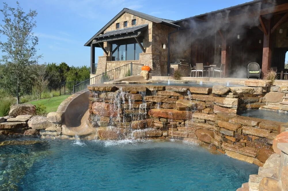 Stacked-stone waterfalls cascading into a freeform pool beneath a stone home with a covered patio