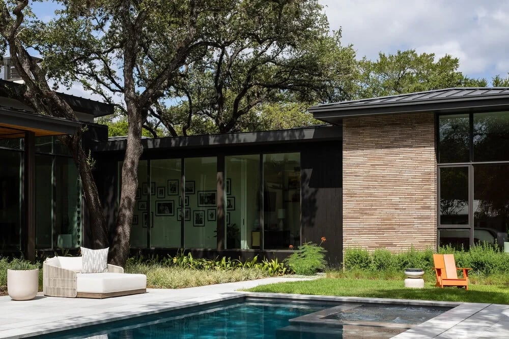 Rectangular pool beside a glass-walled modern home, with lounge seating and orange chairs under oaks
