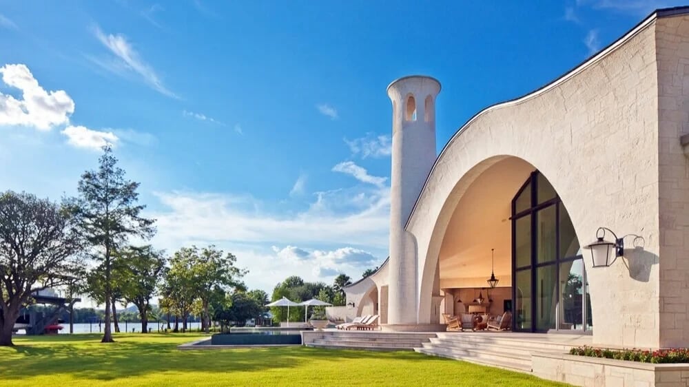 White-stone estate with a great curved arch and round tower above a lakeside lawn and pool