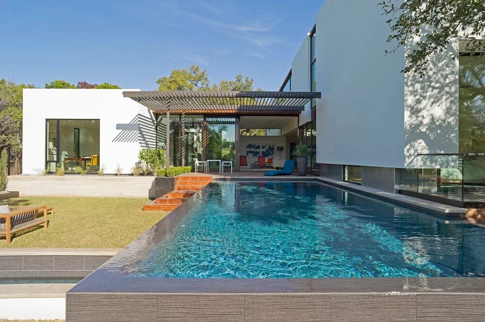 White modern home with a slat pergola beside an infinity pool and lawn under clear skies