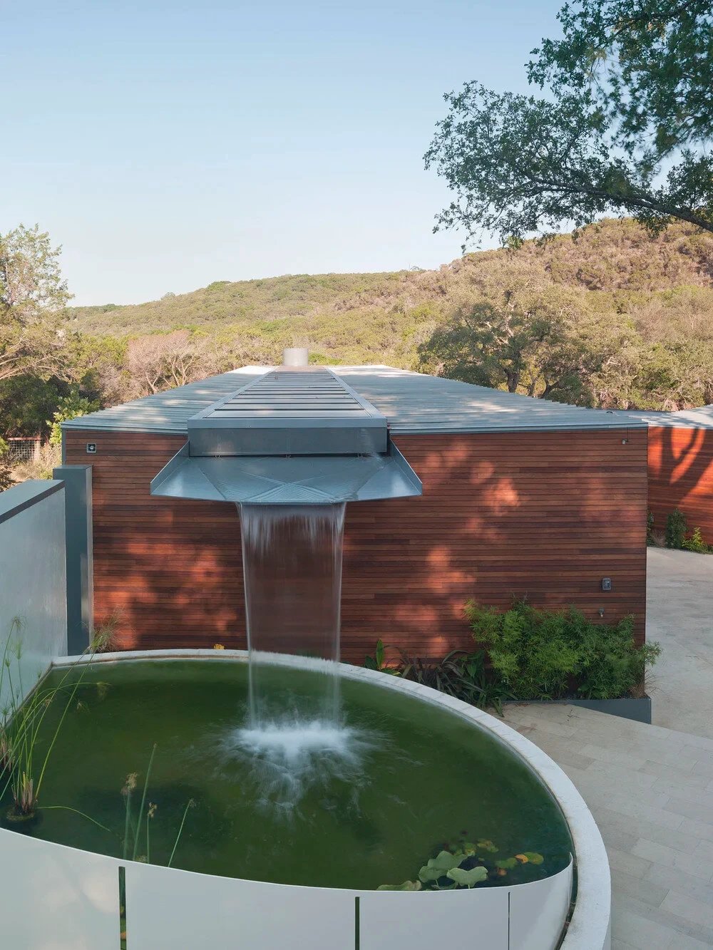 Sheer waterfall spilling off a wood-clad roof into a round stone pond pool, Hill Country beyond