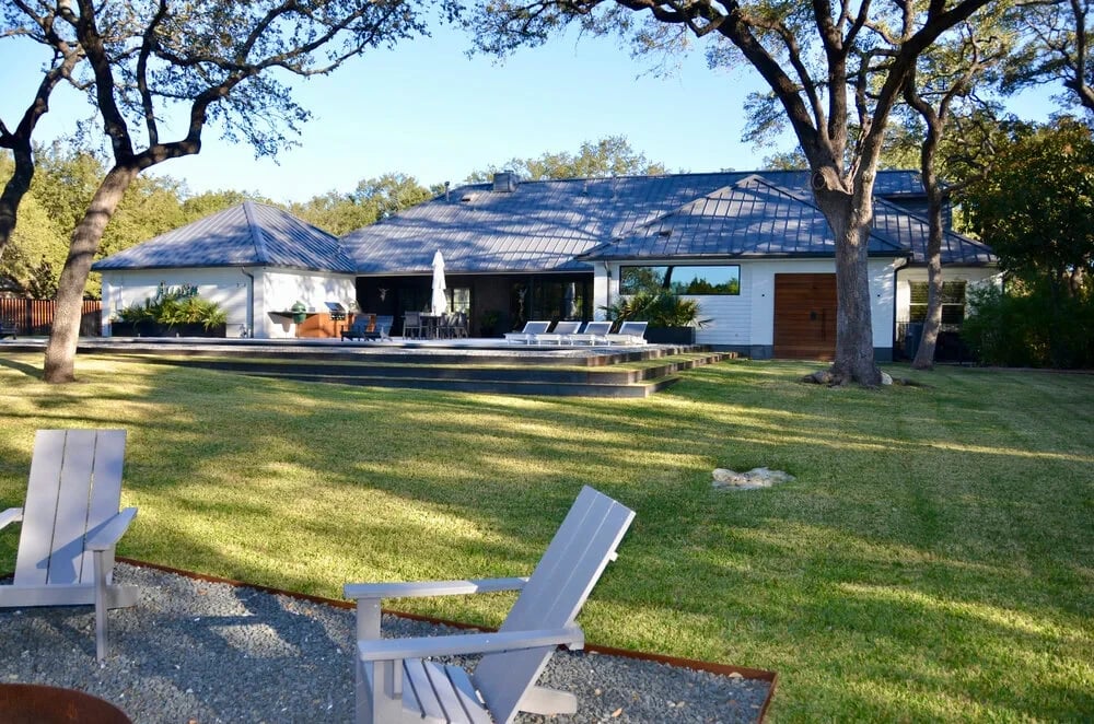 White metal-roof home across a lawn with a pool and lounge chairs, framed by oaks