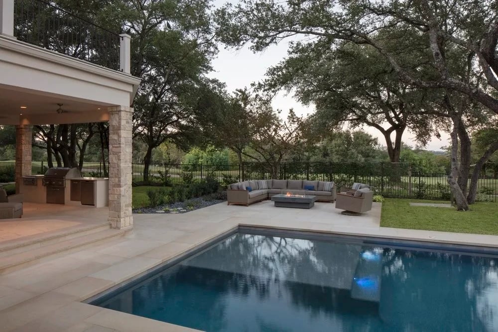 Geometric pool with stone deck and lounge seating below an oak-shaded stone home at dusk