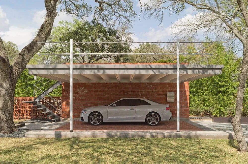 Silver sedan parked under a flat-roofed carport beside a brick-and-glass modern home among trees