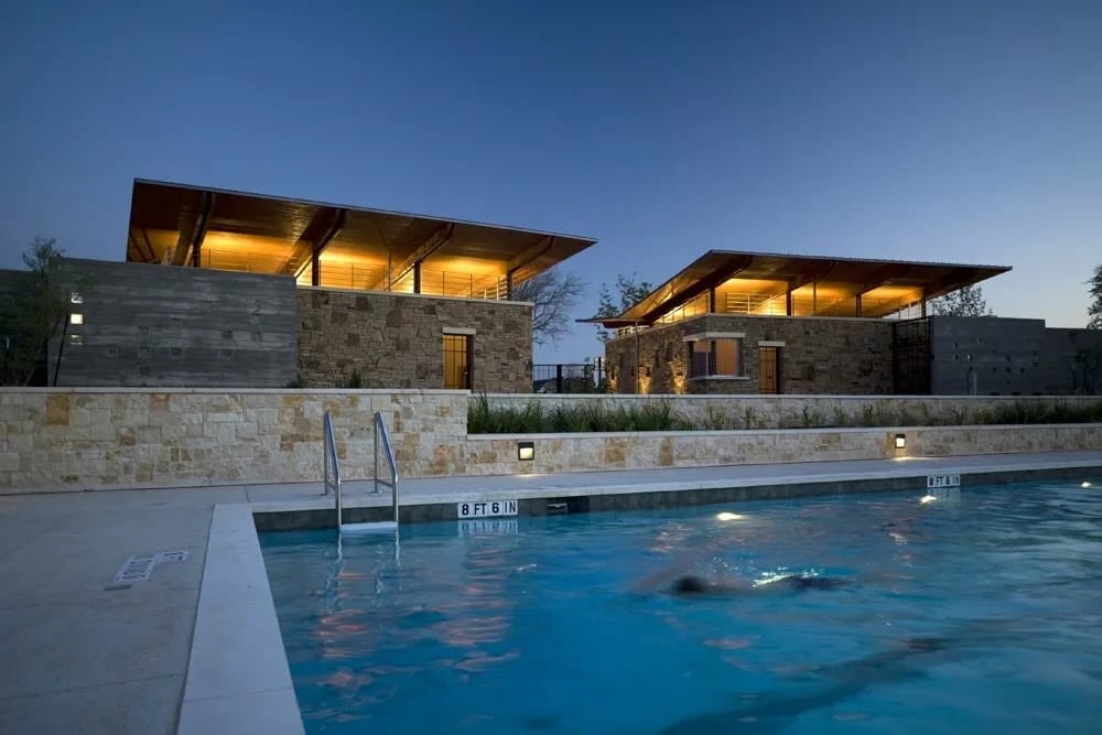 Lit lap pool with a swimmer at dusk below glowing stone-and-timber pavilions with broad roofs