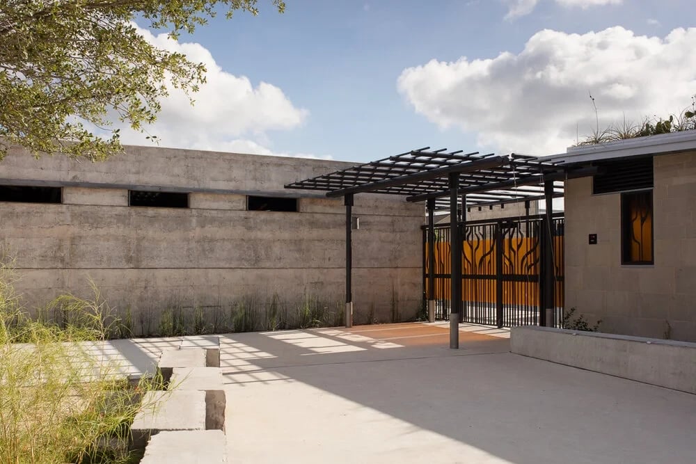 Concrete courtyard with a steel pergola and orange tree-pattern metal gate under blue sky