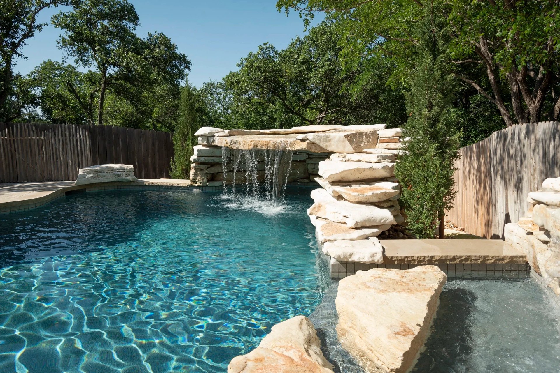 Freeform pool with a clear stone waterfall and rocky spillway edge in a fenced, tree-lined backyard