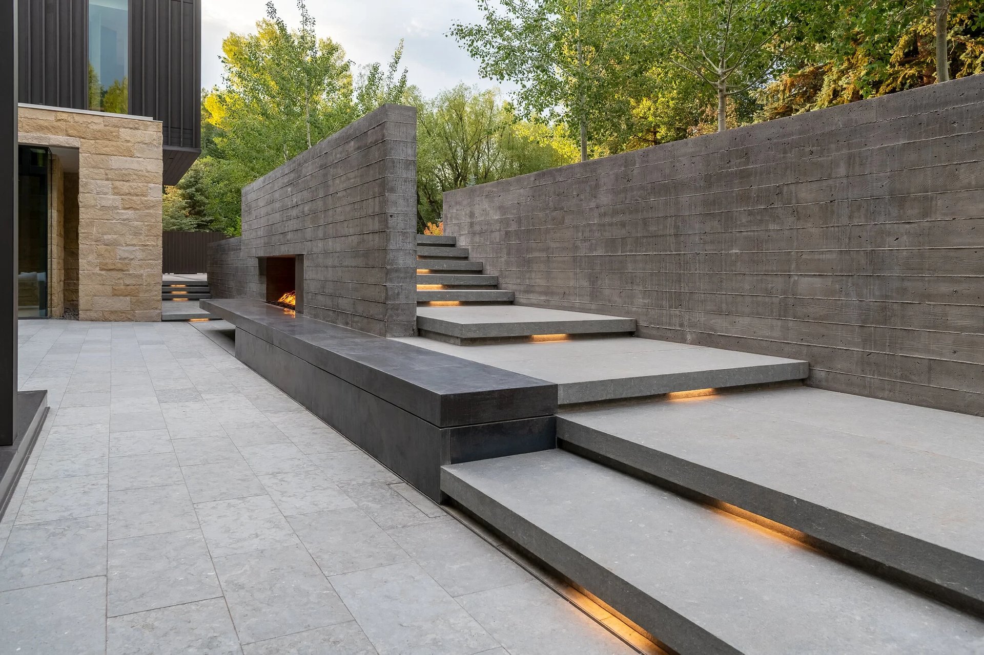 Stone terrace with a recessed fireplace and lit floating steps climbing a board-formed concrete wall