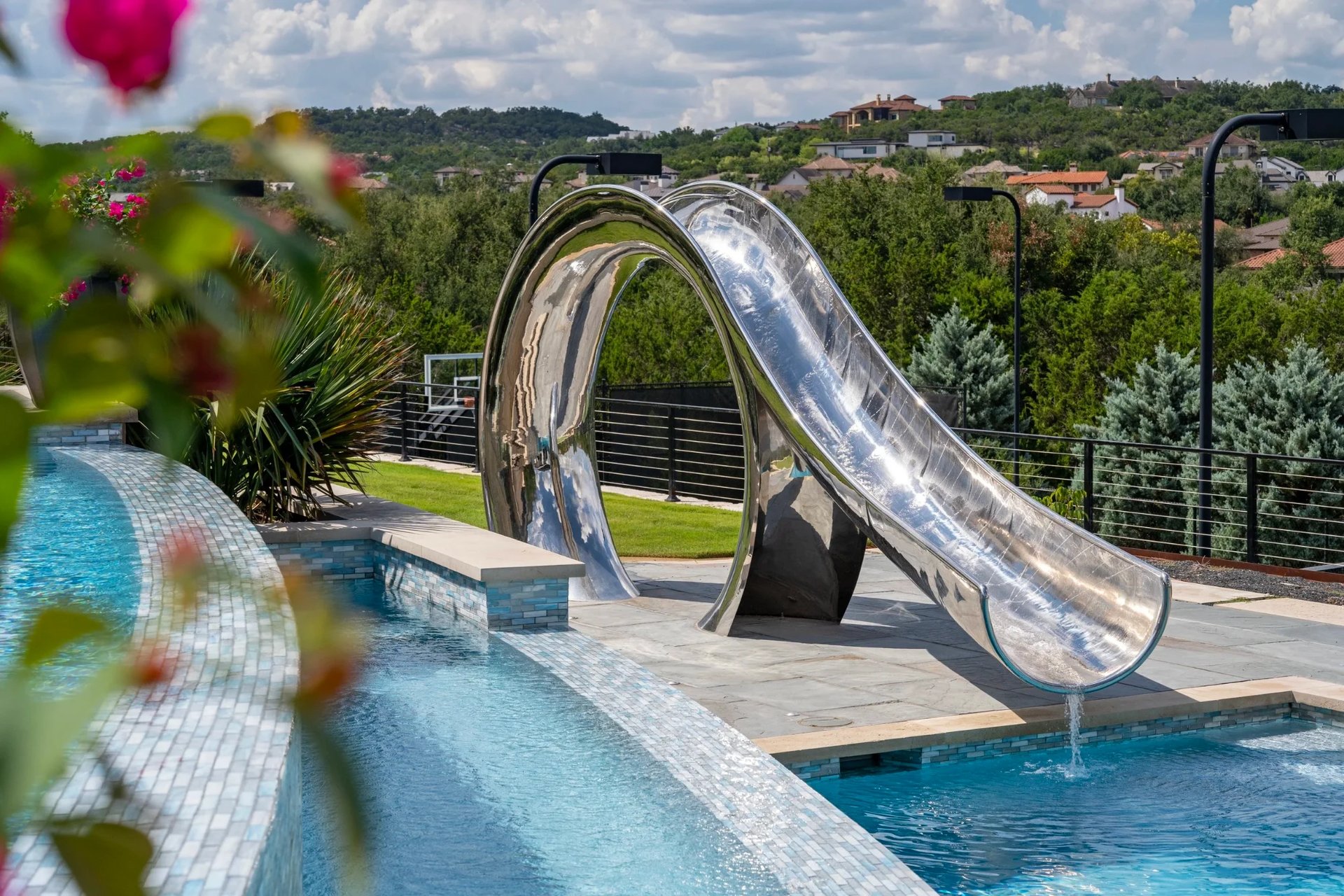 Stainless steel waterslide framed by pink blooms, spilling into a glass-tile pool over a hillside