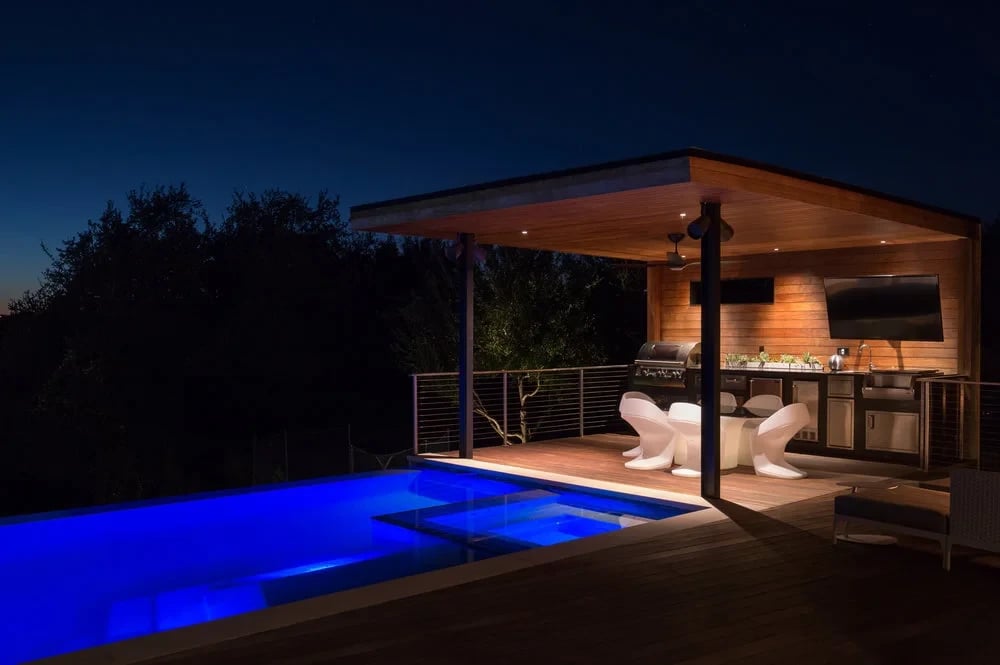 Infinity pool and spa lit blue at night beside a wood cabana with outdoor kitchen and white chairs
