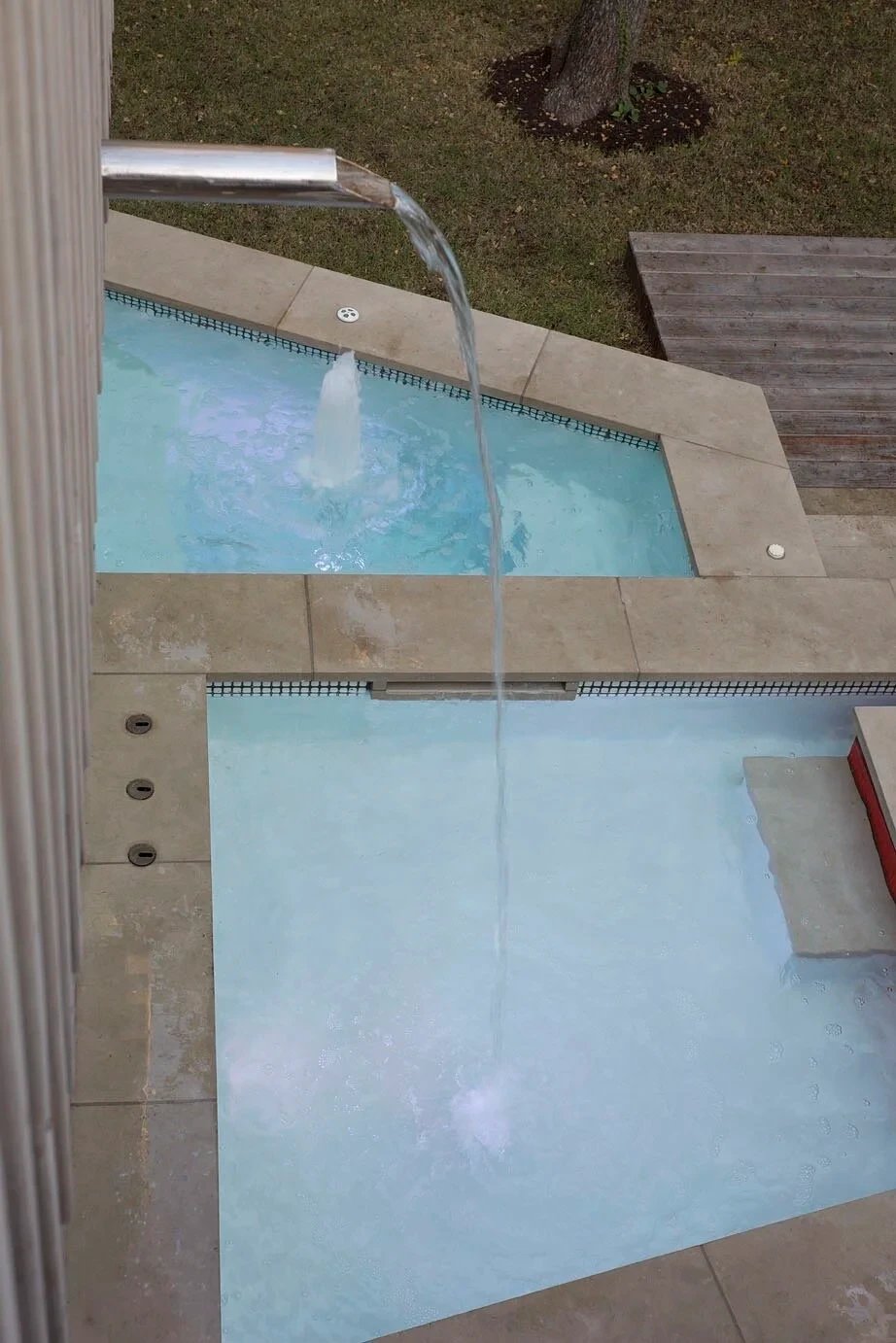 Travertine-edged spa with a scupper spilling into a pale geometric pool below, seen from above