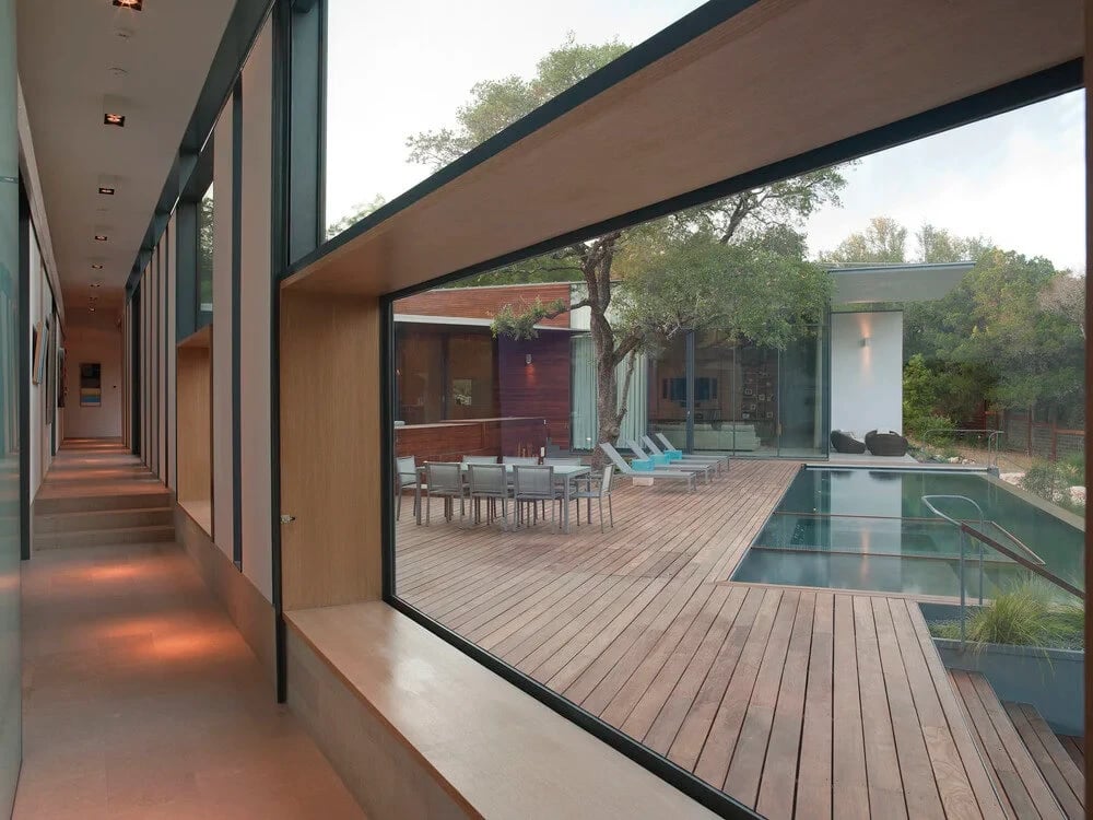 Glass-walled hallway looking out to a wood-deck courtyard, dining set and pool under a live oak