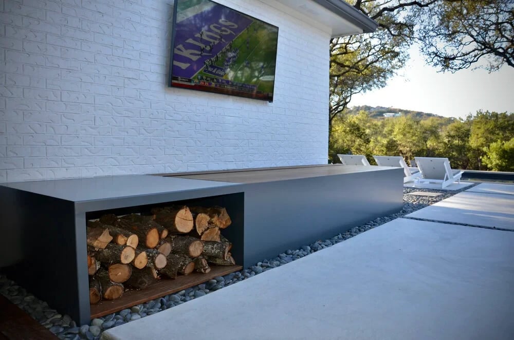 Outdoor lounge counter with stacked firewood below a mounted TV, hills visible beyond