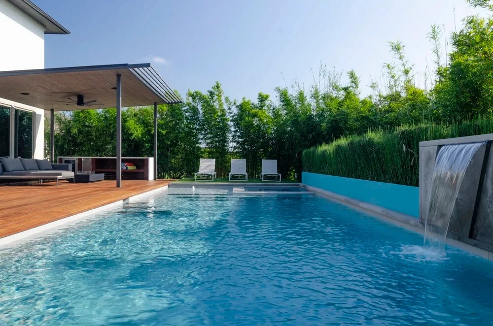 Geometric pool with a sheer-descent spillway and ipe deck framed by bamboo and a covered patio