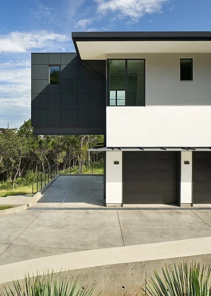 Modern home with a dark cantilevered upper level over a carport, driveway and woods beyond