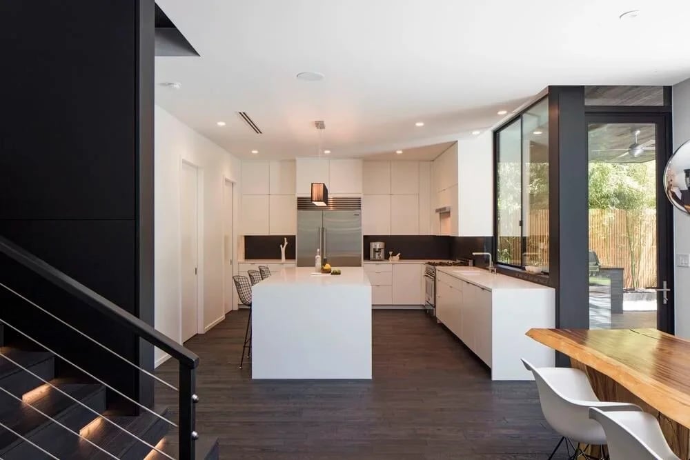 Open white kitchen with a waterfall island, dark backsplash, and stairs leading down to the dining area