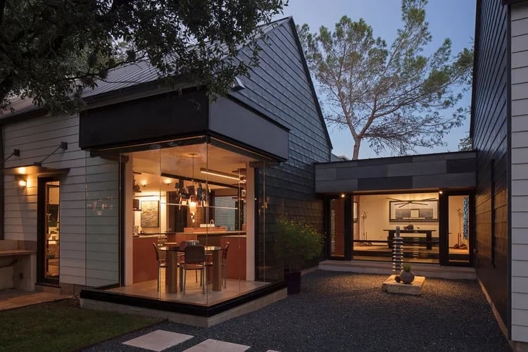 Glass-walled corner of a modern home at twilight with warm interior light, gravel courtyard and pine