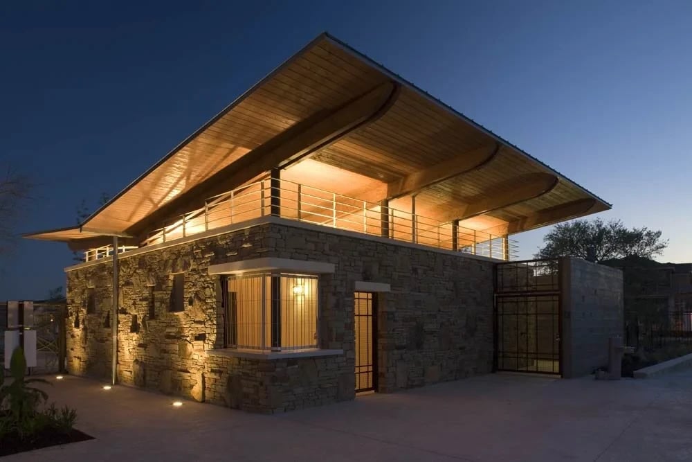 Stone pavilion with a sweeping cantilevered timber roof glowing from within at twilight