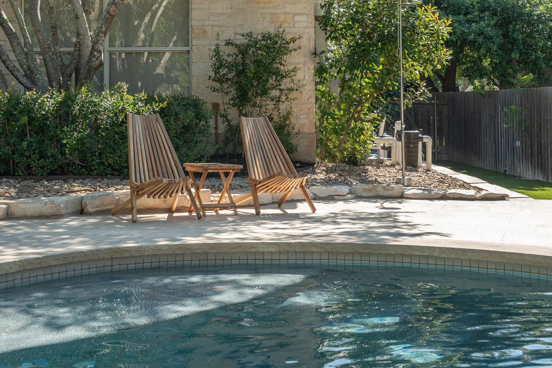 Two teak chairs and a side table on a stone deck beside the rounded edge of a clear pool