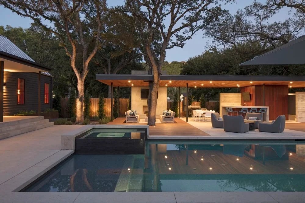Pool and spa glowing at twilight beside a covered patio with lounge seating under tall live oaks