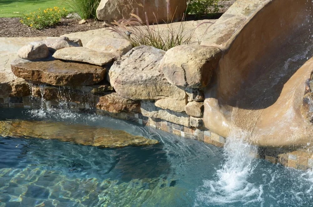 Detail of a freeform pool with a curved stone slide and rock-edged spillway spilling into clear water