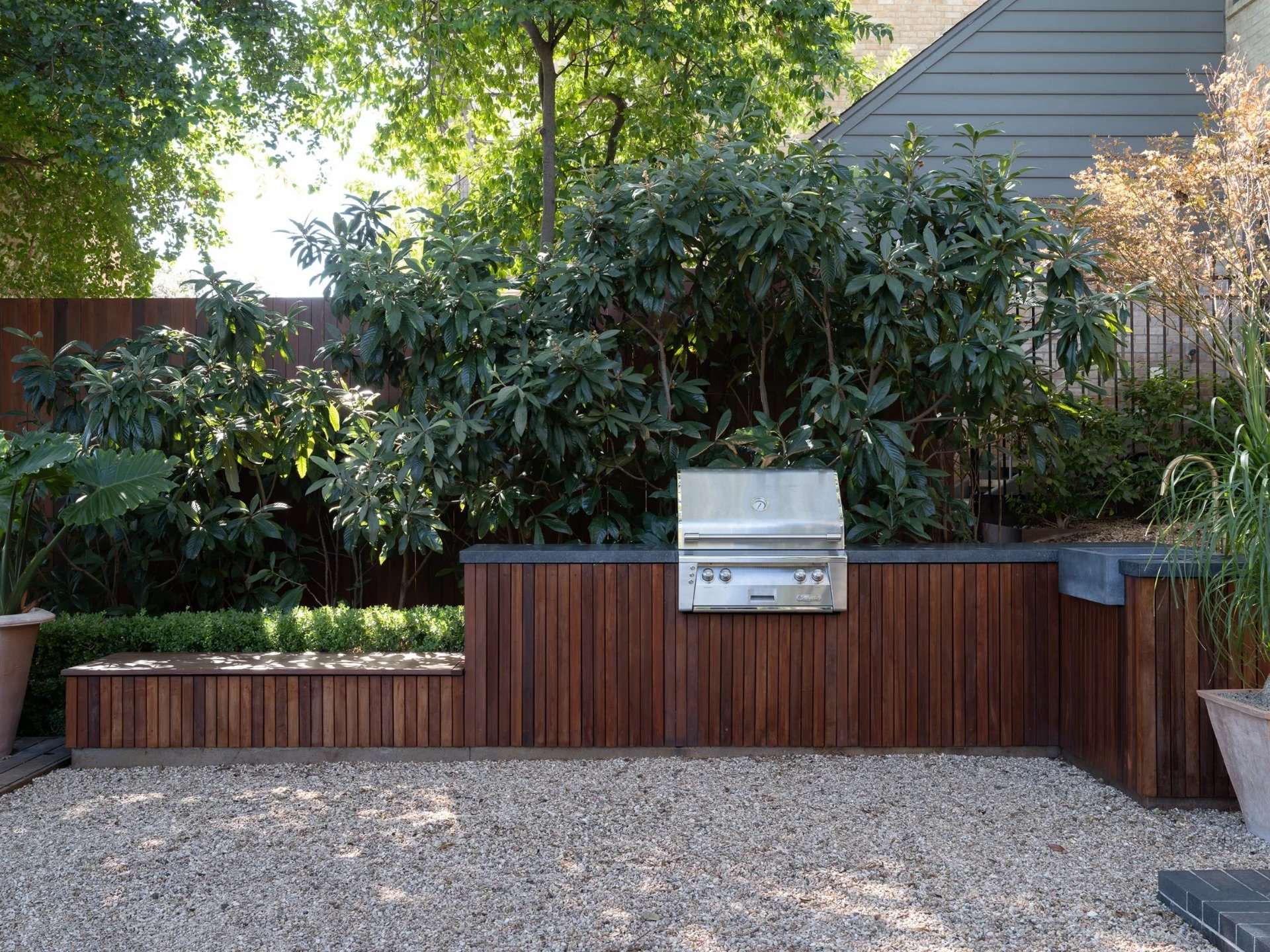 Outdoor kitchen with a built-in grill in a wood-clad counter, set on gravel against tall shrubs