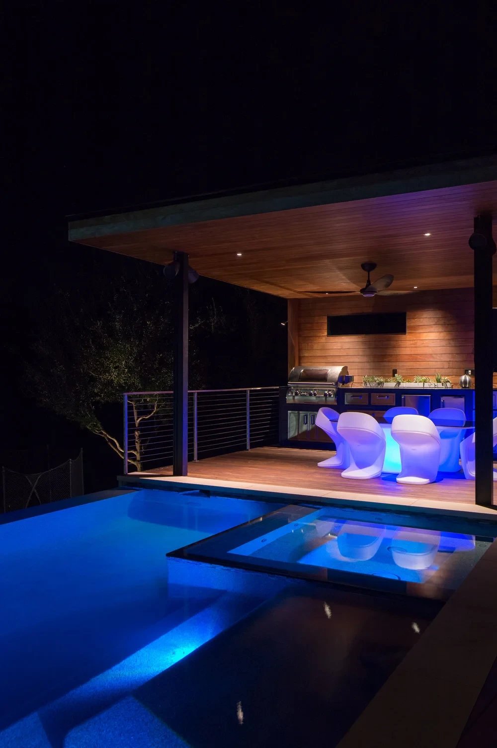 Pool and spa glowing blue at night beside a lit wood cabana with outdoor kitchen and white chairs