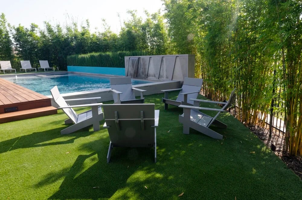 Gray Adirondack chairs on a lawn beside a geometric pool screened by a tall bamboo hedge