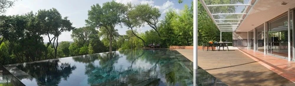 Wide infinity pool reflecting live oaks beside a wood-deck patio under a white pergola