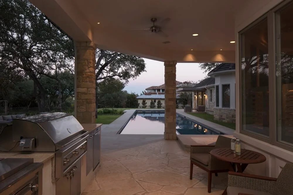 Covered outdoor kitchen with a grill looking out past stone columns to a geometric pool and lawn at dusk