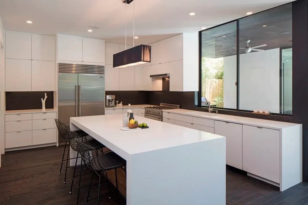 White modern kitchen with a waterfall-edge island, wire bar stools, and glass wall to a covered patio