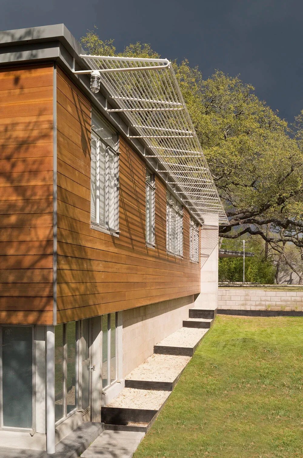 Cedar-clad home wall with a white steel sunshade and steps down a green lawn under a dark stormy sky