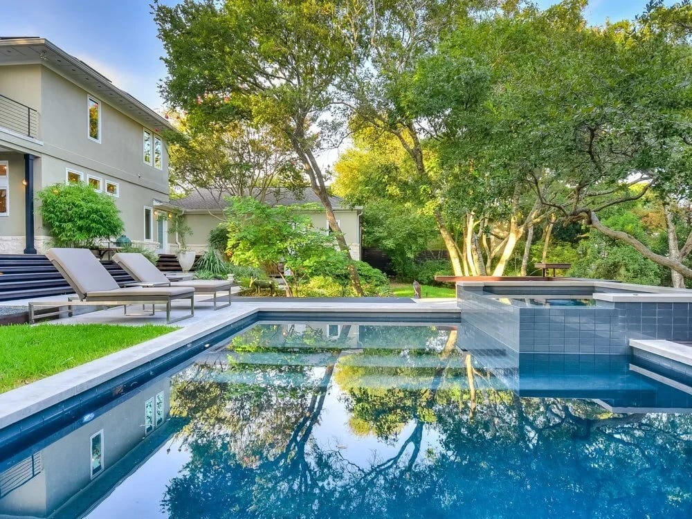Geometric pool reflecting a home and trees in late light, loungers lined up on the stone deck