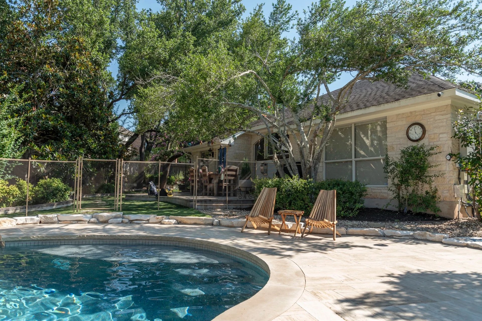 Single-story stone home with shade trees and teak chairs beside a curved pool on a travertine deck