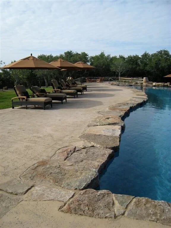 Freeform pool with a stacked-stone edge, flagstone deck, umbrellas, and chaise lounges