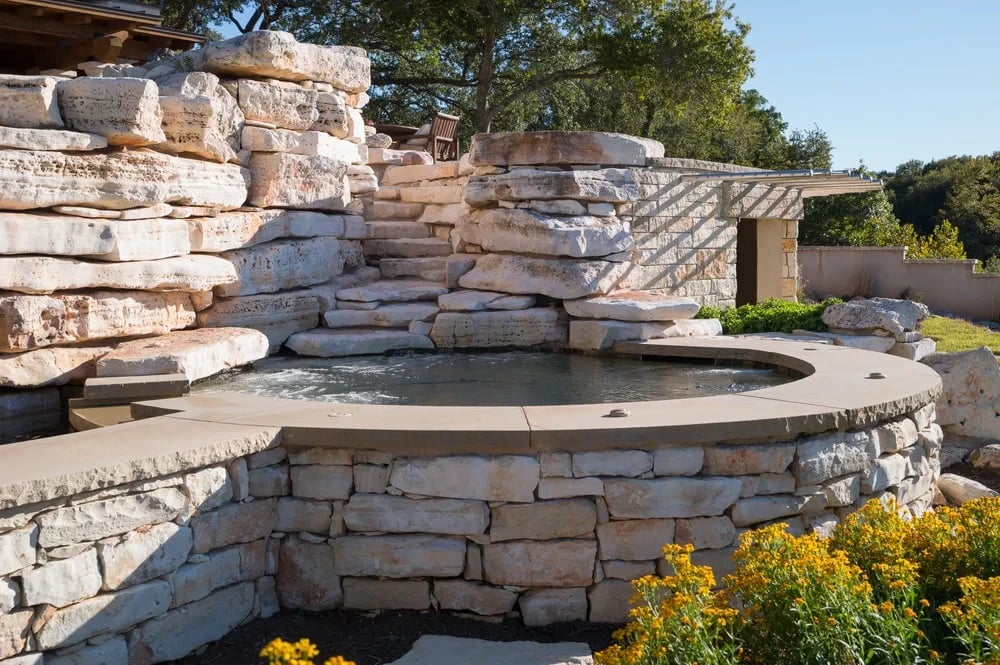 Round stone spa with stacked-stone waterfall surround and flagstone deck among native plantings