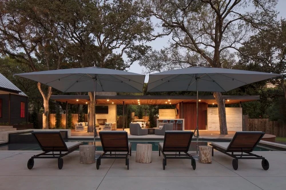 Cantilever umbrellas over lounge chairs on a patio at dusk, an outdoor kitchen lit beyond